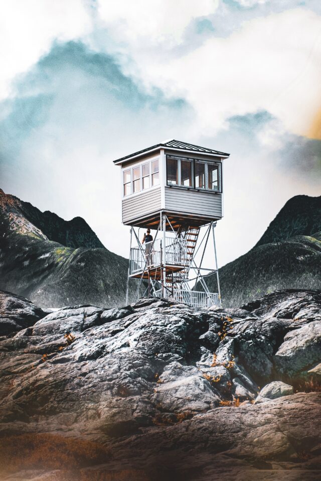 An old watchtower on top of a rocky ridge, with tall forested mountains and a cloudy sky in the background.